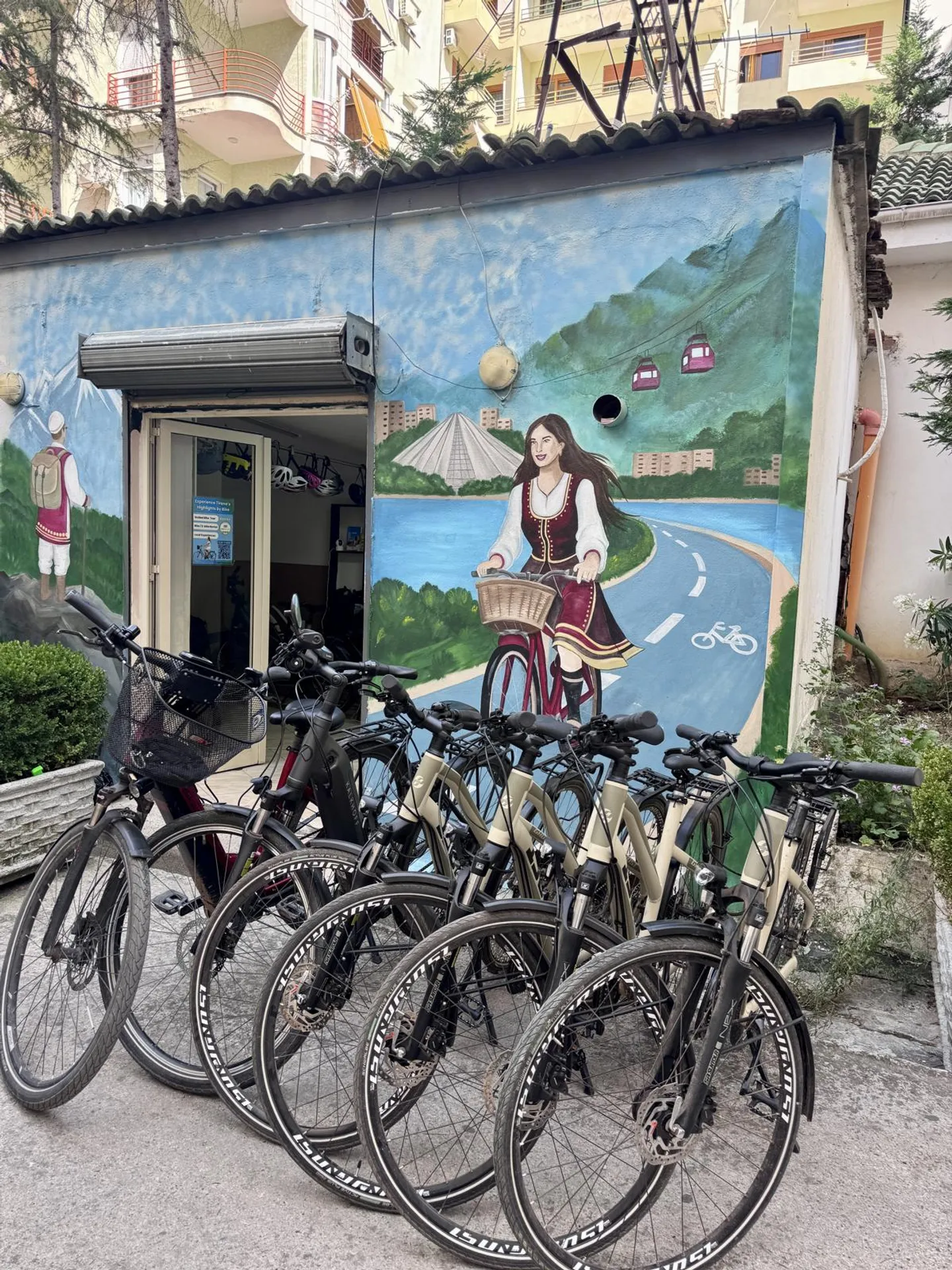 A row of bicycles parked in front of the shop, decorated with a mural of a person in traditional Albanian clothing