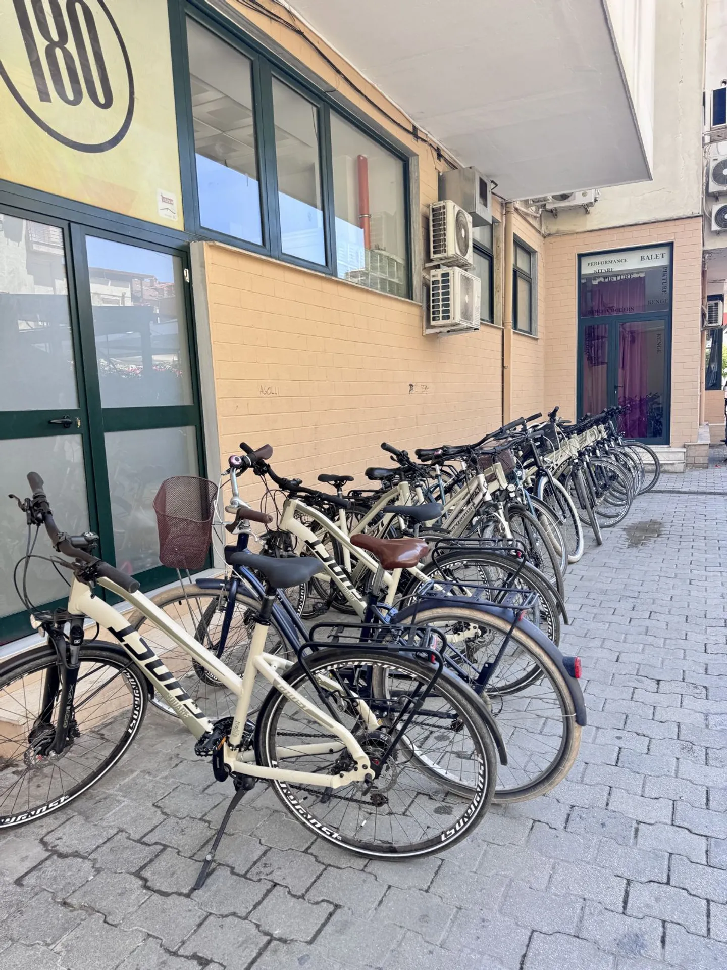 Një rresht biçikletash bezhë me qira të parkuara në trotuar pranë dyqanit BikeTirana